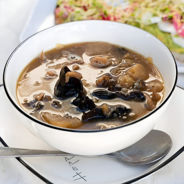 A white bowl filled with a dark broth soup containing mushrooms and other vegetables.