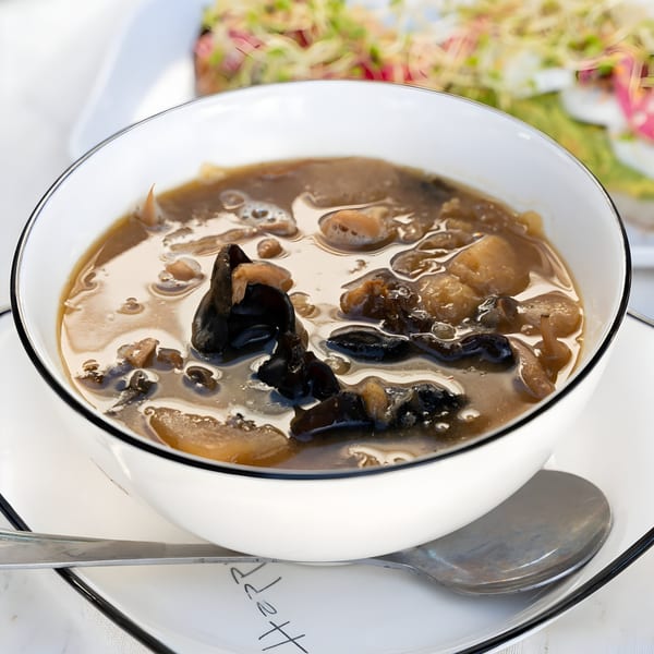 A white bowl filled with a dark broth soup, black mushrooms, and other ingredients, with a spoon.