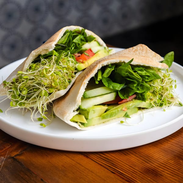 Two healthy pita sandwiches filled with fresh greens, sprouts, avocado, cucumber, and tomato.