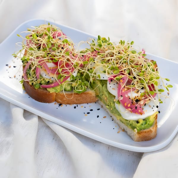 Two avocado toasts topped with sliced eggs, pickled onions, sprouts, and everything bagel seasoning.