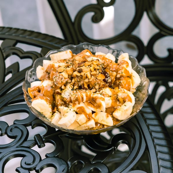 A glass bowl of sliced bananas topped with peanut butter, nuts, and seeds on a black ornate table.
